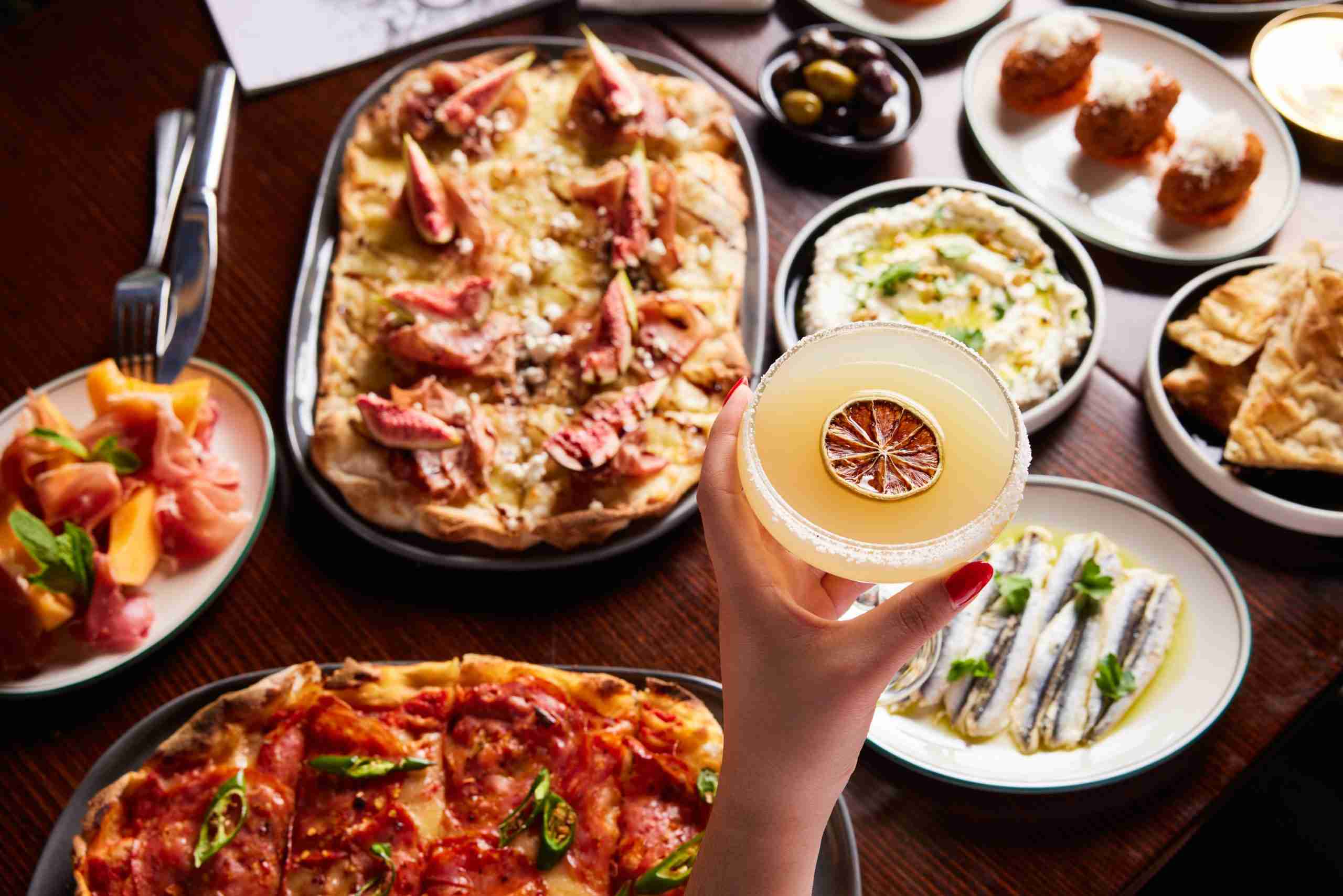 Margarita cocktail with pinsa pizza, marinated anchovies and other summer menu items in the background.