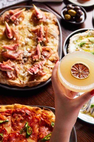 Margarita cocktail with pinsa pizza, marinated anchovies and other summer menu items in the background.